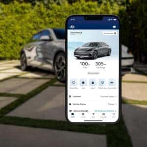 A smartphone displaying the Hyundai Bluelink app with vehicle status, range, and controls, with a 2025 Hyundai IONIQ 6 parked in the background.