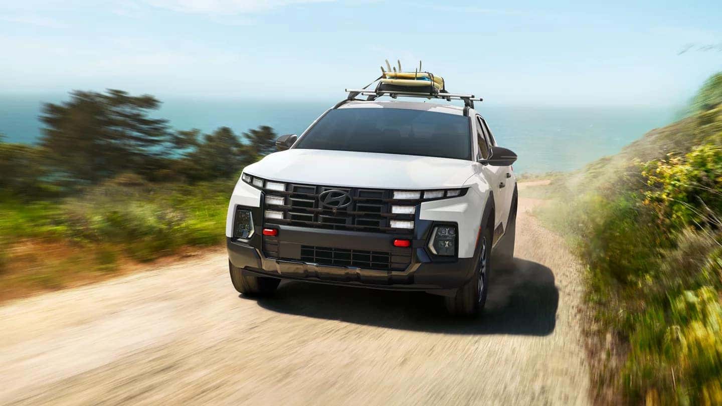 a white santa cruz driving on a gravel path near the ocean with surfboards on the roof rail