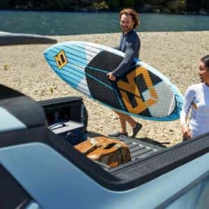 a woman standing next to a hyundai santa cruz looking at a man with a surf board standing on an ocean beach