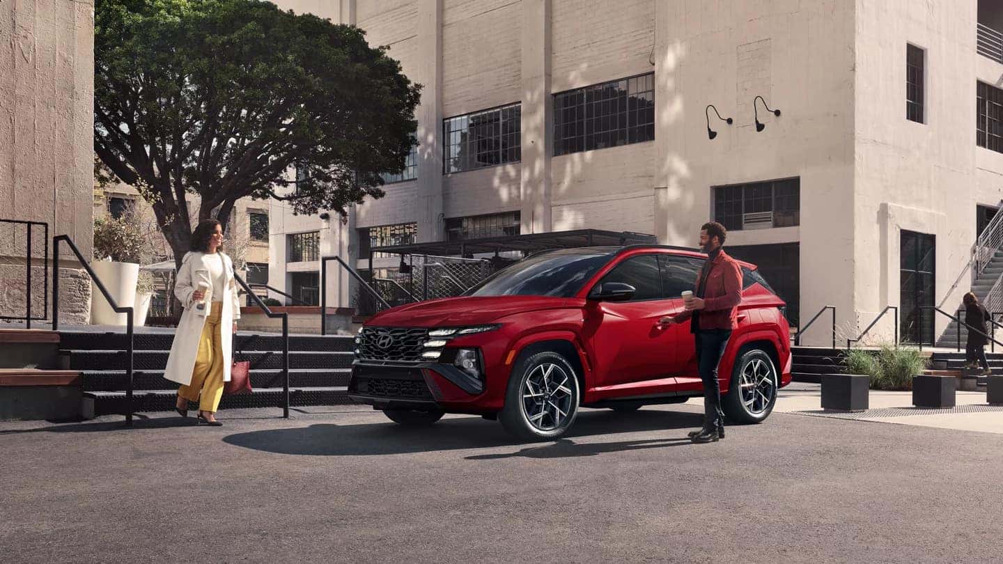 A red 2025 Hyundai Tucson parked in front of a white industrial building with two people approaching it - a woman in white coat and yellow pants and a man in business attire.