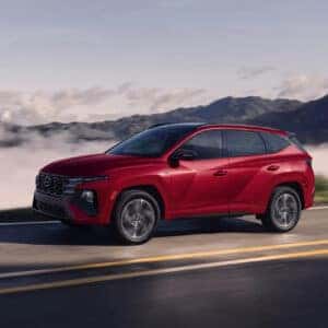 Dynamic shot of a red 2025 Hyundai Tucson driving on a mountain road with fog in the valley below, showcasing its sleek profile and aerodynamic design.