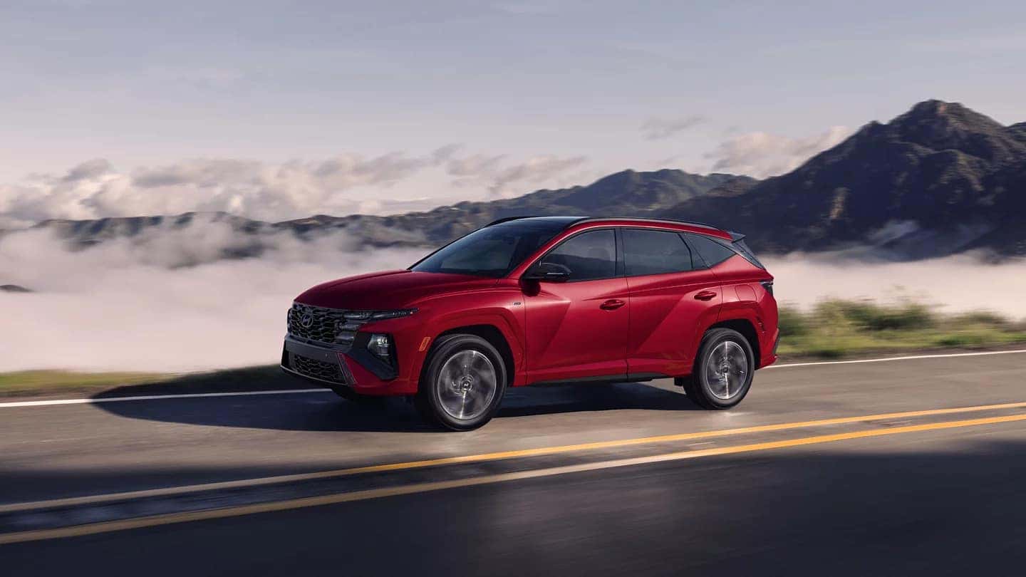 Dynamic shot of a red 2025 Hyundai Tucson driving on a mountain road with fog in the valley below, showcasing its sleek profile and aerodynamic design.