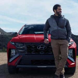 A smiling couple standing in front of their red 2025 Hyundai Tucson in a mountain setting, dressed in outdoor adventure clothing.