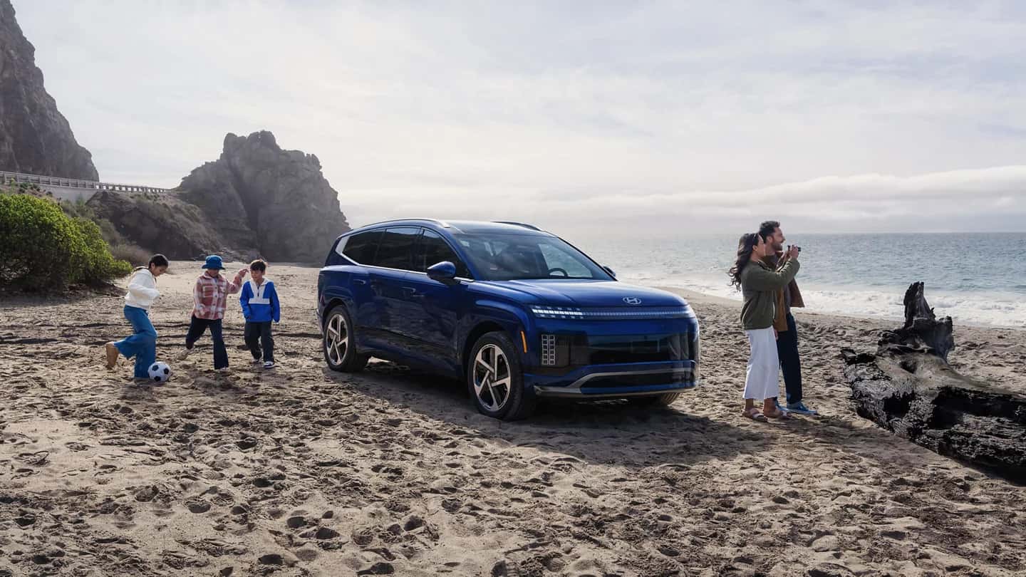 a blue 2026 ioniq 9 on the beach near the ocean with a couple taking photos and three children playing soccer