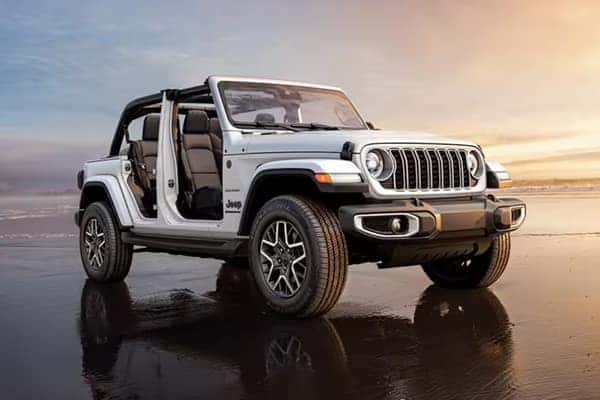 White 2025 Jeep Wrangler, doors and top removed, parked on a beach at sunset, emphasizing open-air freedom.